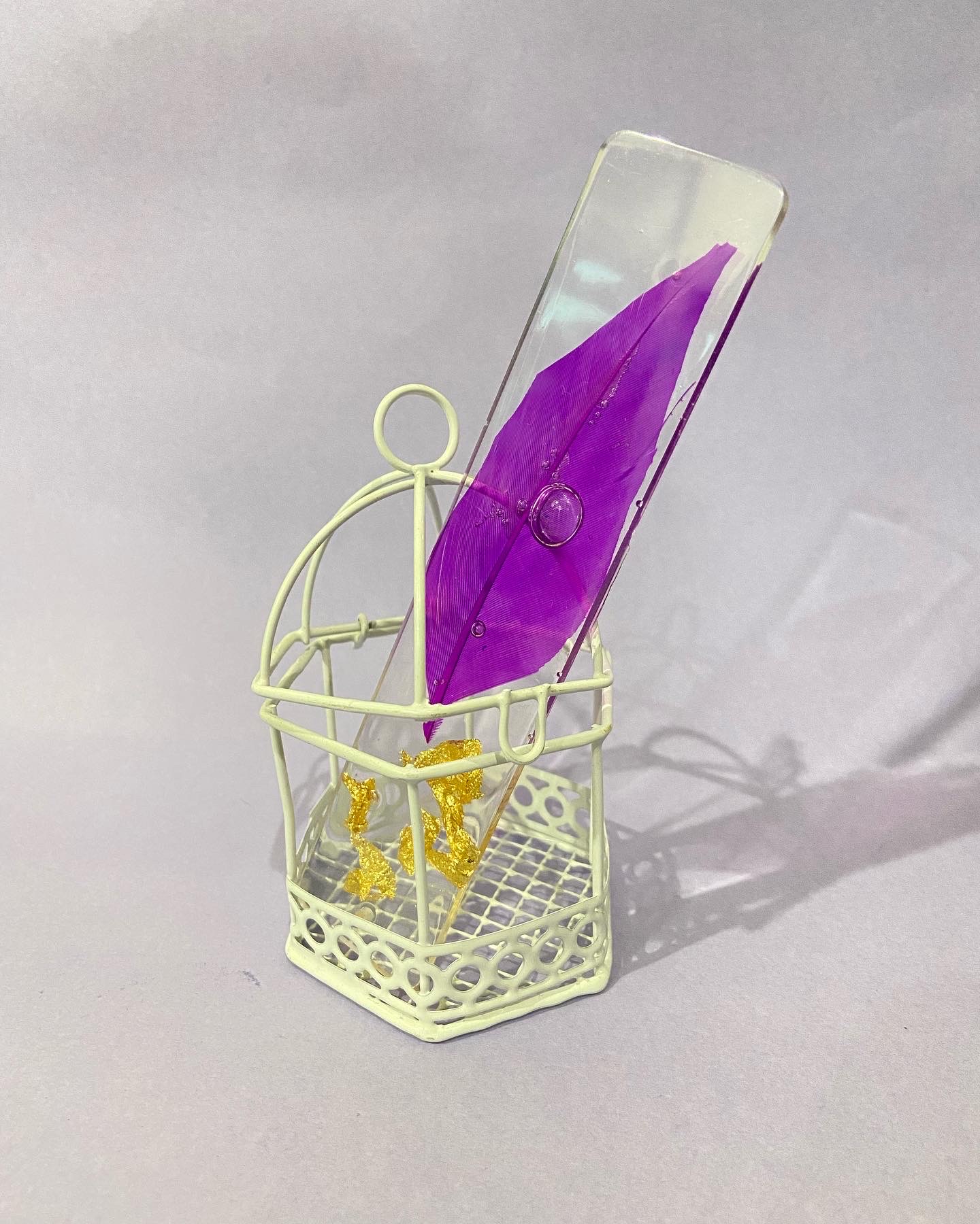 Resin bookmark with a purple feather and gold flakes displayed in a mint birdcage